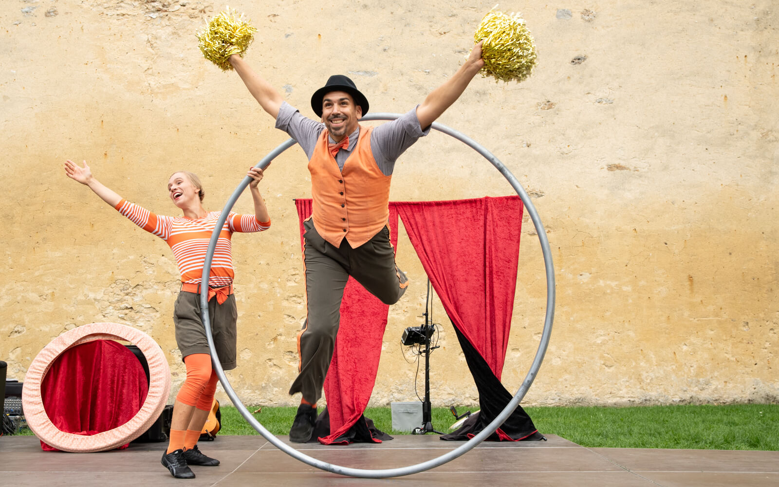 Circus artists: woman raising arms, man in a Cyr wheel with golden pompoms.