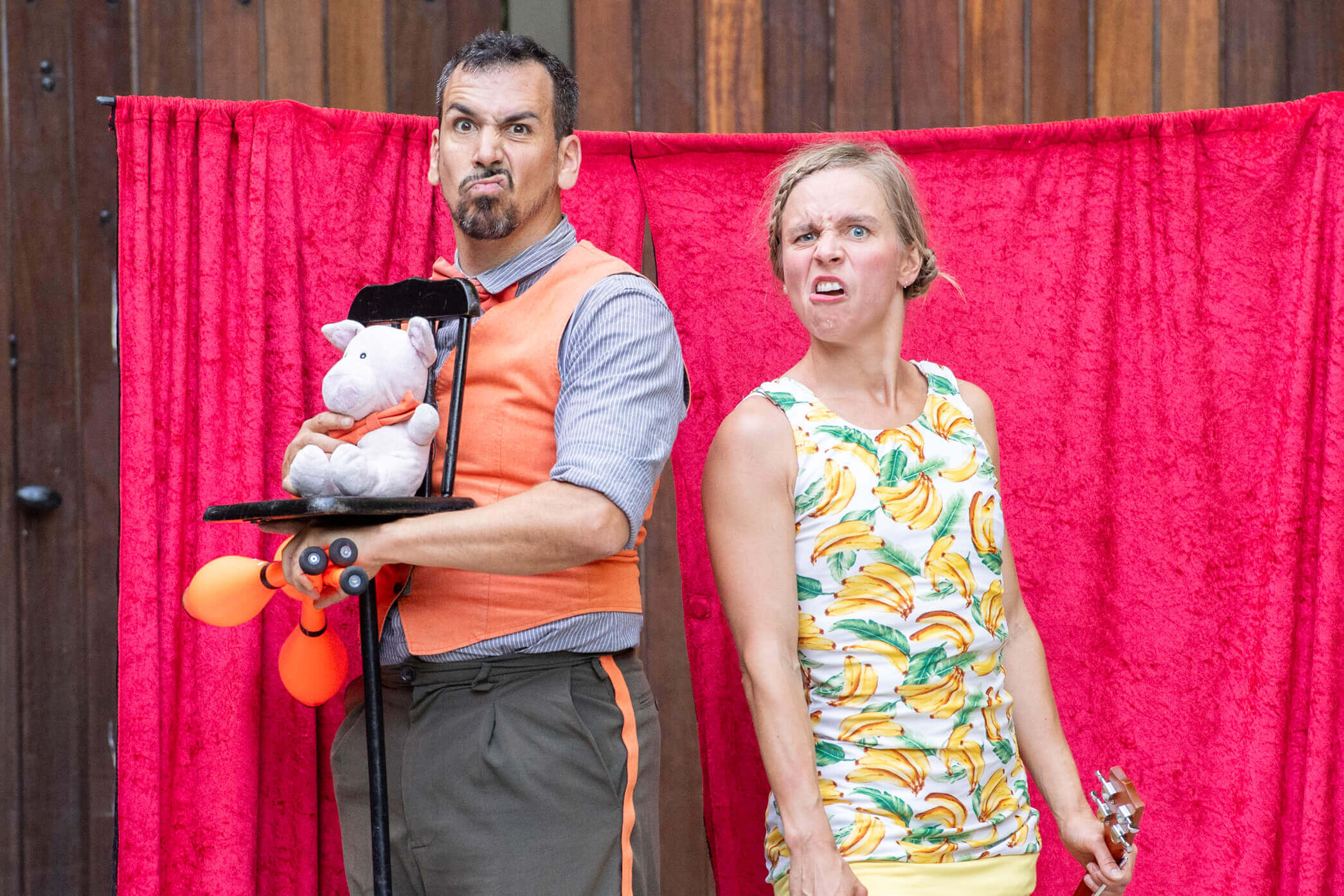 Male artist with pig puppet, female artist with surprised expression, outdoor circus performance, red curtain.