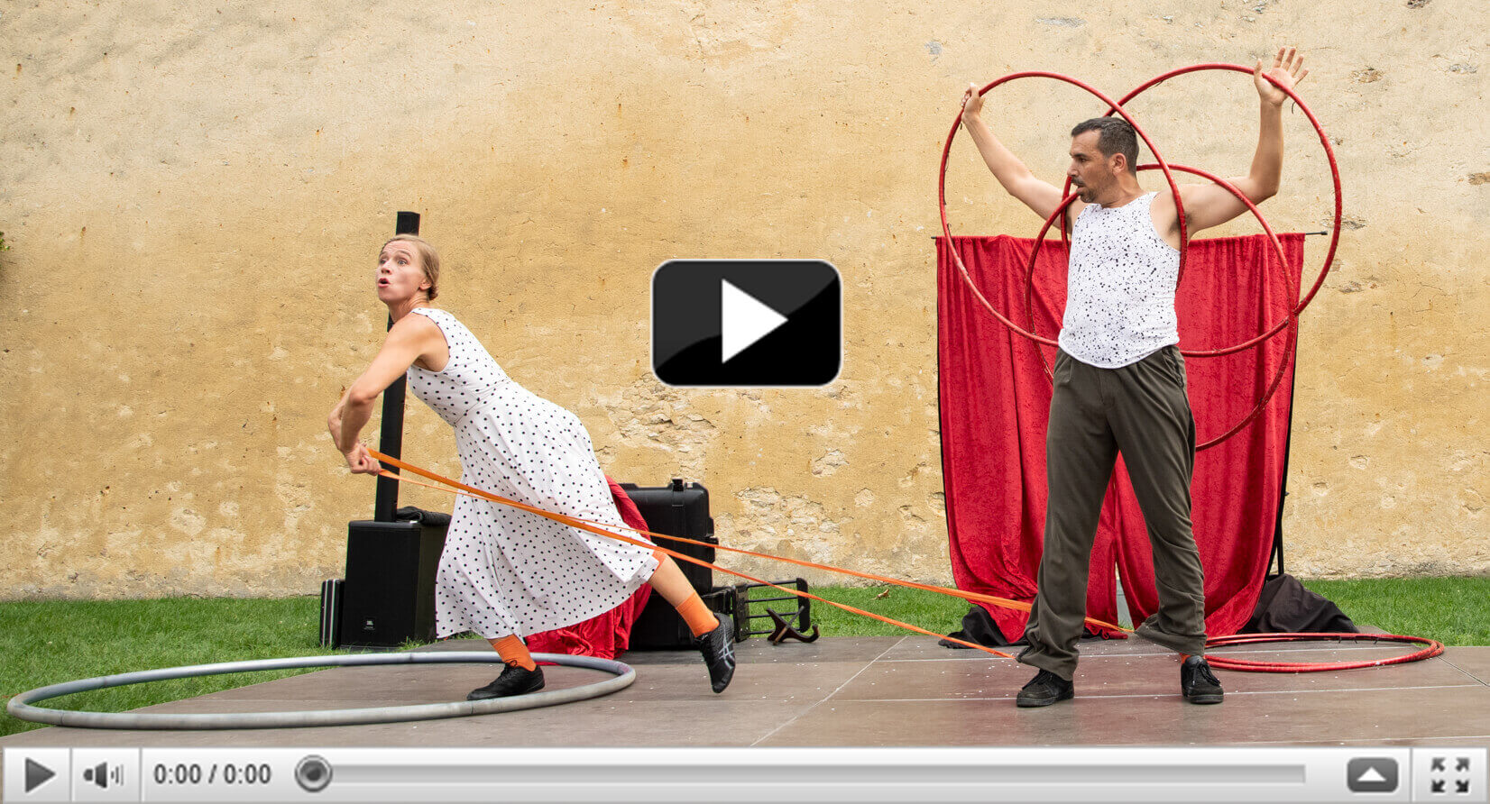 Woman in polka dot dress, man in white shirt forming a heart with two hula hoops, street arts and circus performance, beige wall in the background.