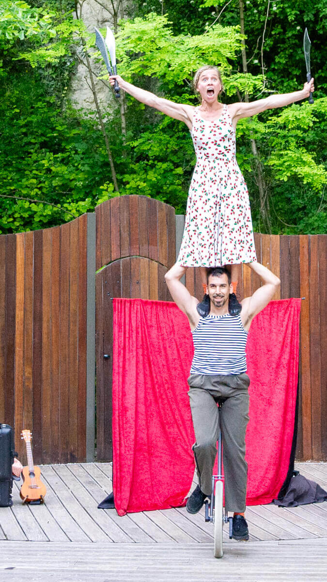 Artiste du Cirque Les Dudes juchée sur les épaules d'un collègue, jonglant avec des couteaux durant le spectacle Stories in the City! sur une scène extérieure en bois.